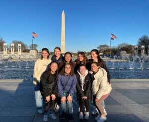 Nine girls standing together in front of the Washington Monument in Washington, DC, under a clear blue sky with two American flags on either side in the background.