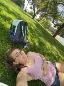 Me, in a pink striped top and jean shorts, lying down on a grassy hill with a teal backpack behind me.